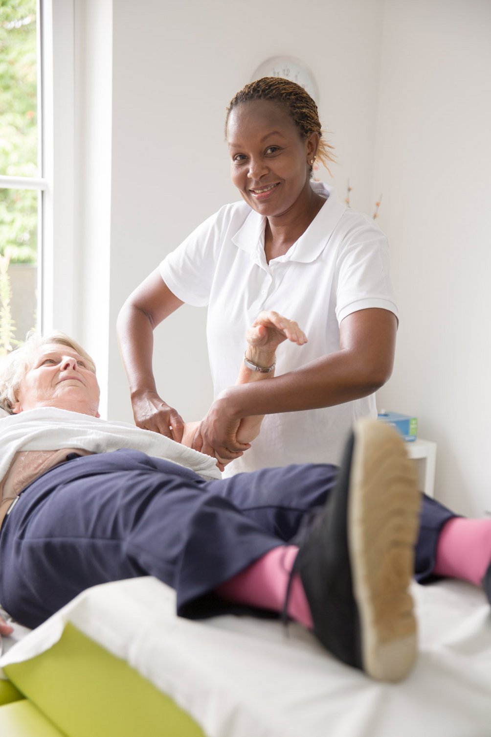Die manuelle Therapie hilft, Funktionsstörungen des Bewegungsapparates zu beheben. Therapeutin unterstützt lächelnd das angehobene Bein einer liegenden Patientin.
